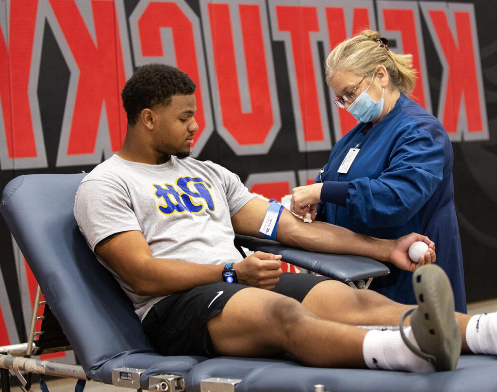 A healthcare worker prepares to draw blood from a seated man in a sports facility.